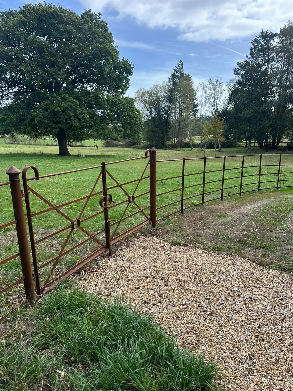 Pair of oxide-brown X-braced estate gates with ring details, set into curved estate fencing beside a gravel track and horse paddock